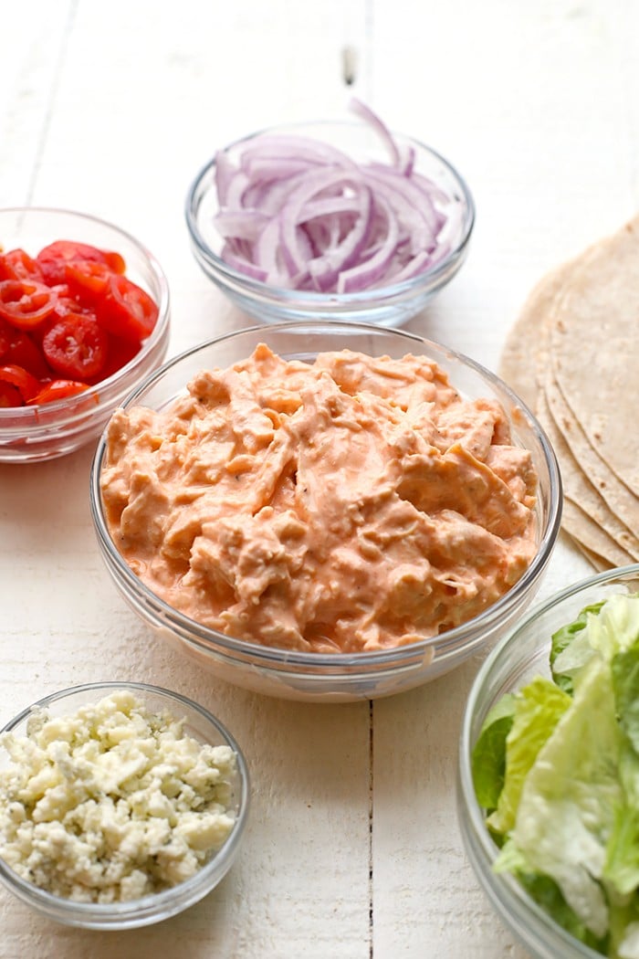 chicken wrap recipe ingredients in individual bowls including tomato, onion, lettuce and buffalo chicken
