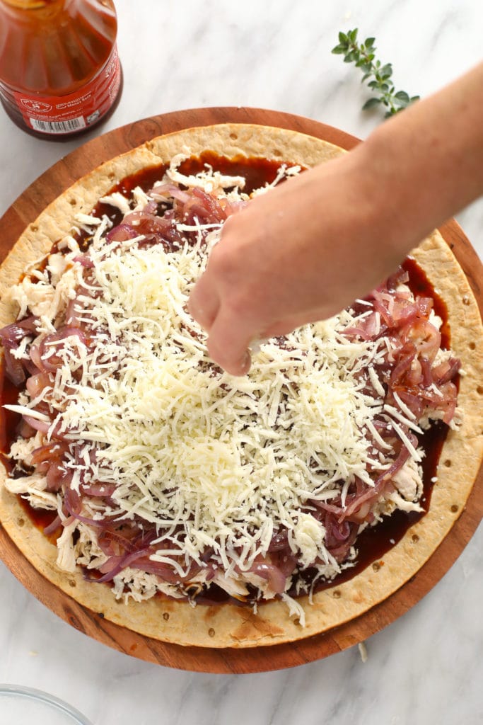 This BBQ chicken pizza with caramelized onions is ready in under 30 minutes and you only need 5 ingredients. It is saucy, flavorful, and makes Friday night pizza night as easy as can be! sprinkling cheese on pizza