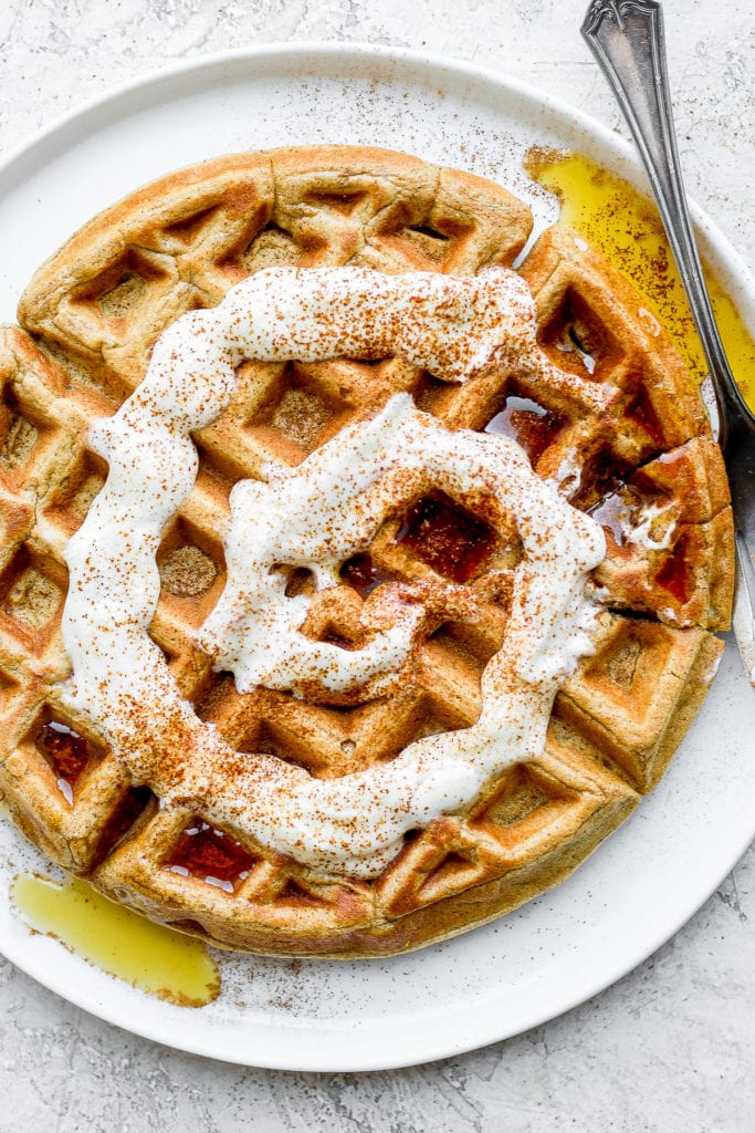 It's a waffle morning over here! These Cinnamon Roll Waffles taste just like the real thing, but pack healthy simple ingredients. Finish with a Greek yogurt cream cheese drizzle for the most delicious breakfast ever! cinnamon roll waffle on a plate with maple syrup and a cream cheese drizzle
