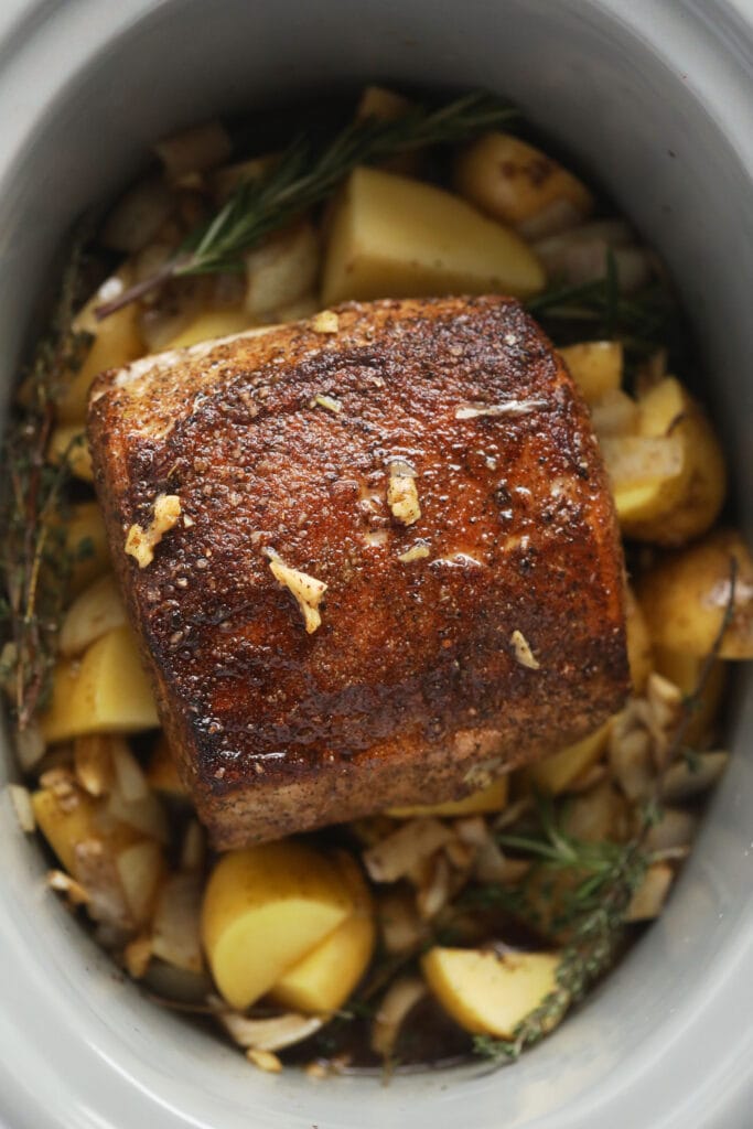 This is the most tender Slow Cooker Pork Loin roast recipe ever! It's served with potatoes and a savory gravy for the perfect meal. Enjoy! slow cooker pork loin roast on a bed of potatoes and herbs in the slow cooker