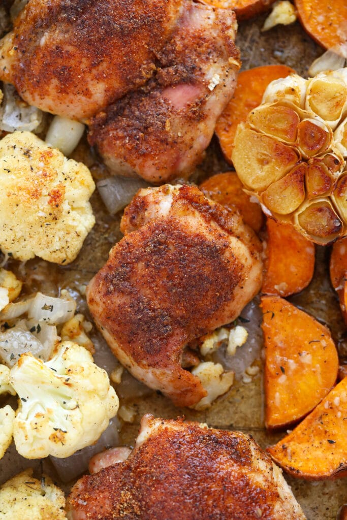 Baked chicken thighs, roasted garlic, cauliflower, and sweet potatoes on a pan.