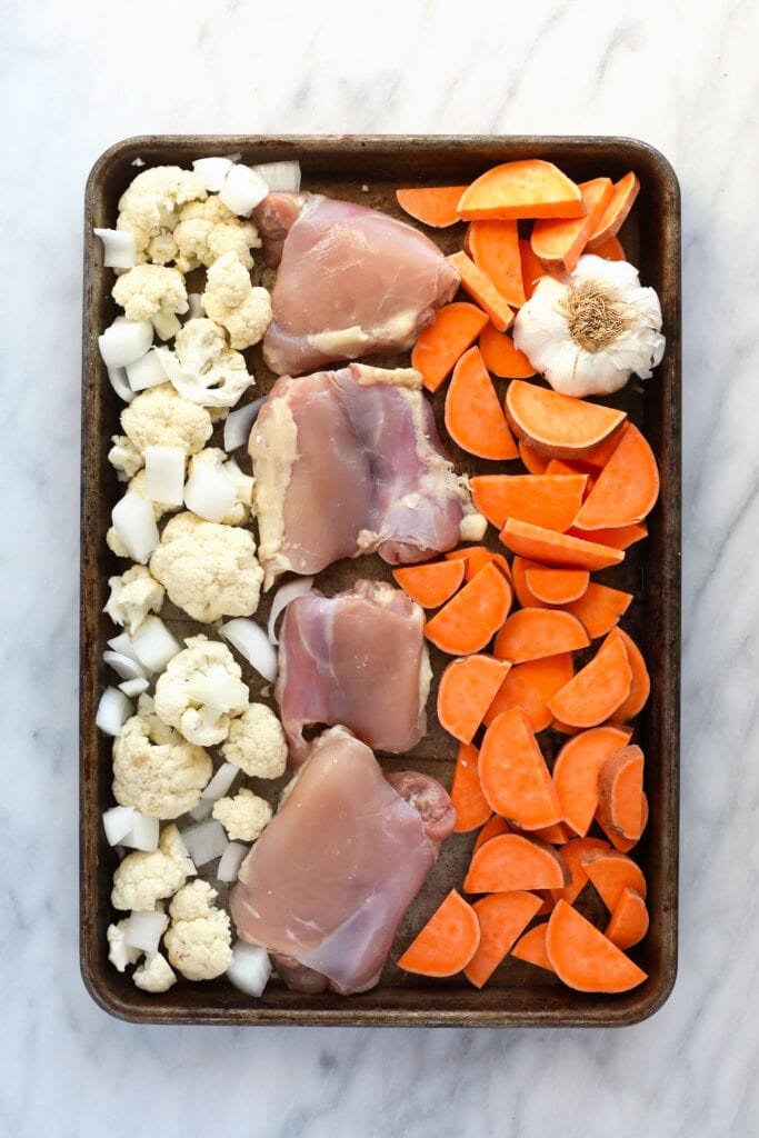 Raw chicken, cauliflower, sweet potatoes, onion, and garlic on a baking sheet ready to bake.
