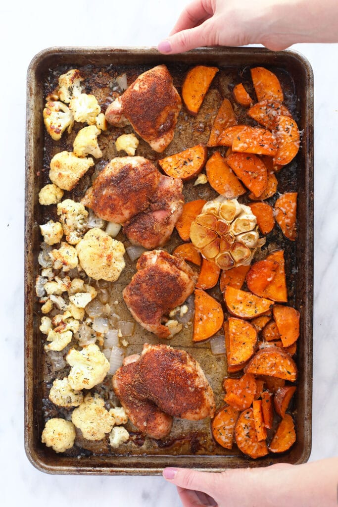 Chicken thighs, sweet potatoes, cauliflower, and garlic on a baking sheet.