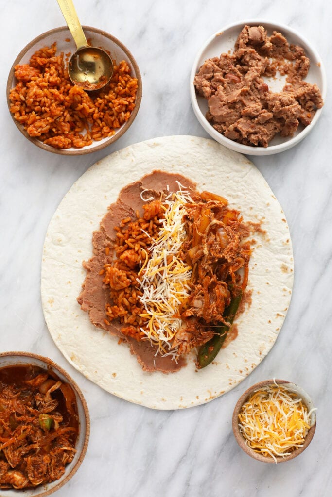 These easy shredded chicken burritos are super flavorful with smothered chicken and fajita veggies, refried beans and Spanish rice. Make a batch and enjoy immediately, or follow our freezer instructions for the ultimate meal prep recipe! shredded chicken burrito ingredients being assembled onto a flour tortilla
