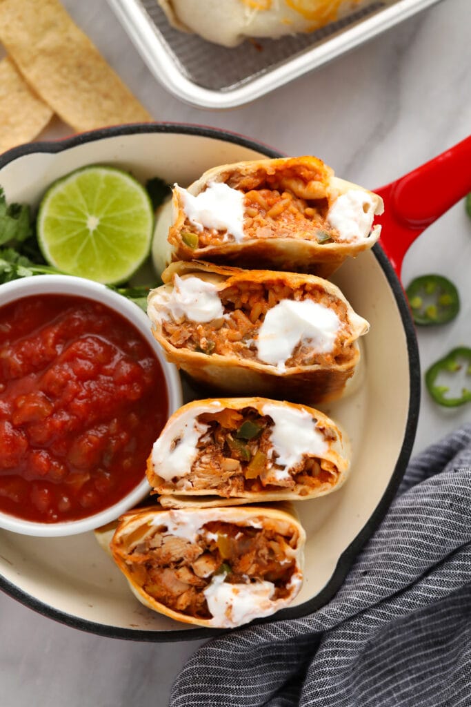 These easy shredded chicken burritos are super flavorful with smothered chicken and fajita veggies, refried beans and Spanish rice. Make a batch and enjoy immediately, or follow our freezer instructions for the ultimate meal prep recipe! shredded chicken burritos cut in half and displayed with a bowl of salsa