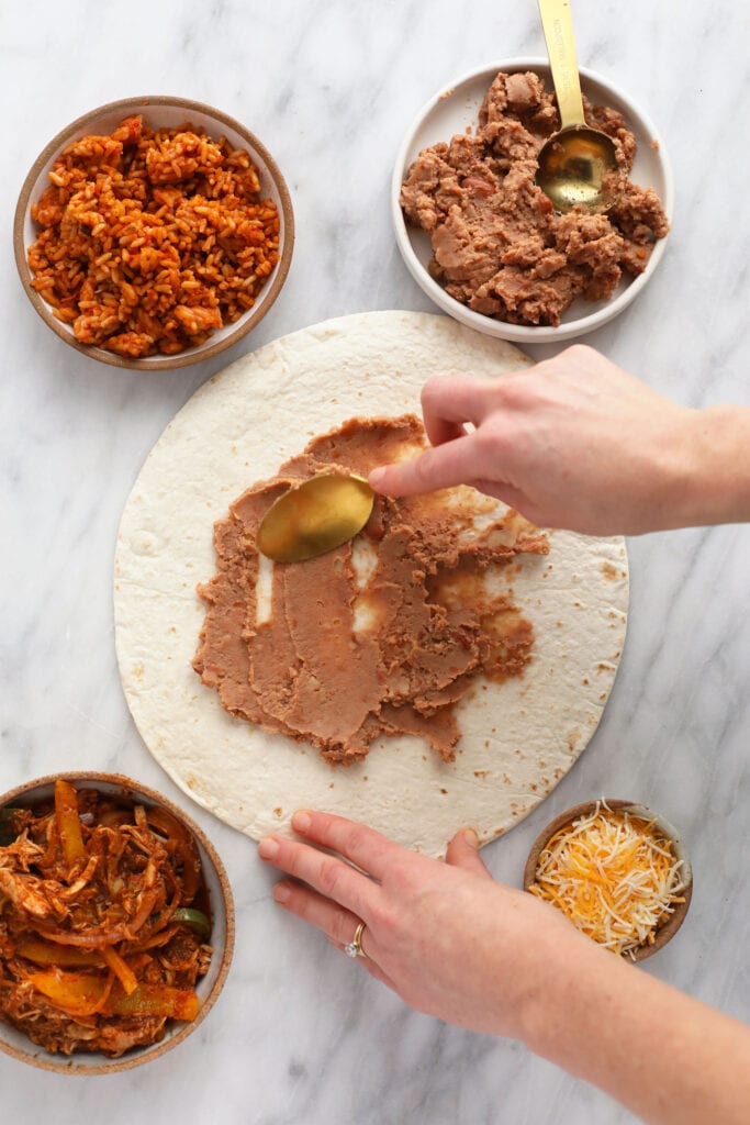 These easy shredded chicken burritos are super flavorful with smothered chicken and fajita veggies, refried beans and Spanish rice. Make a batch and enjoy immediately, or follow our freezer instructions for the ultimate meal prep recipe! refried beans being spread on a tortilla for shredded chicken burritos