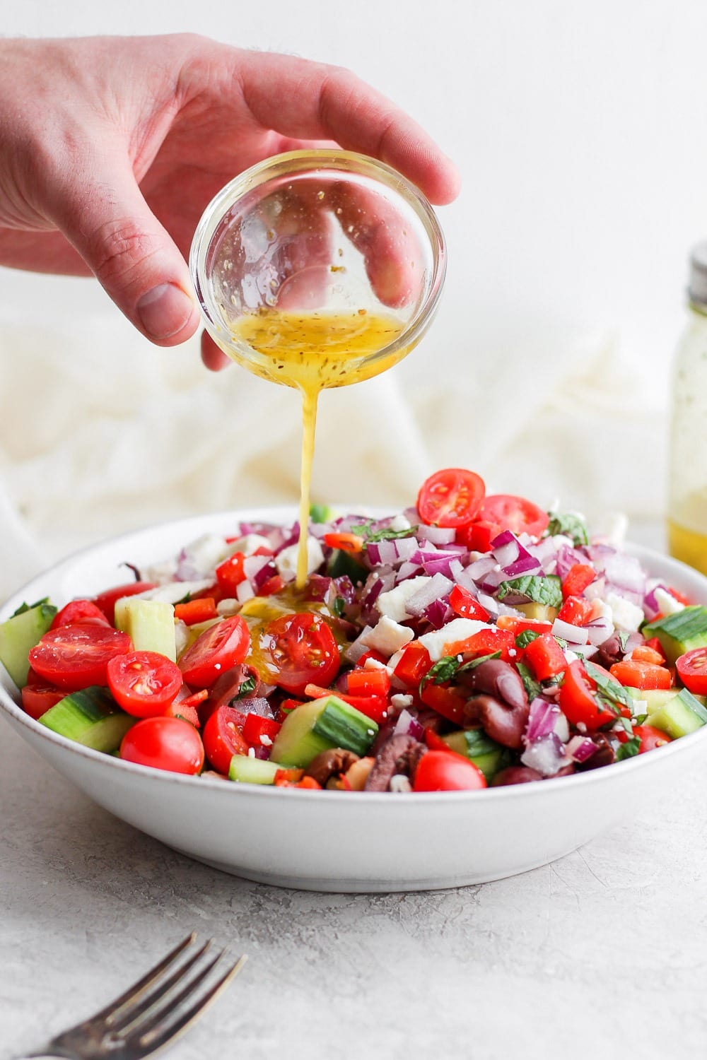 This Greek salad is made with sweet cherry tomatoes, cucumber, red onion, feta, and homemade Greek salad dressing! It is perfect for meal prep, a summer picnic, or just a healthy summer meal! Pouring dressing onto the Greek Salad