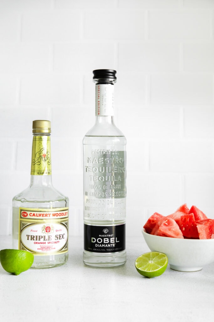 Triple Sec, tequila, fresh watermelon and lime wedges on a counter top.