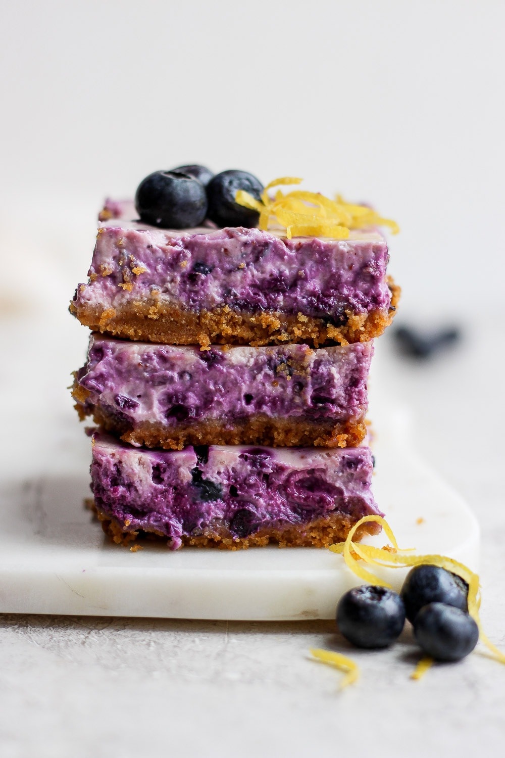 These Blueberry Cheesecake Bars are the perfect summer treat! Equal parts tart and sweet, and paired with a delicious graham cracker crust. Enjoy! blueberry cheesecake bars stacked three high looking delicious and ready to eat
