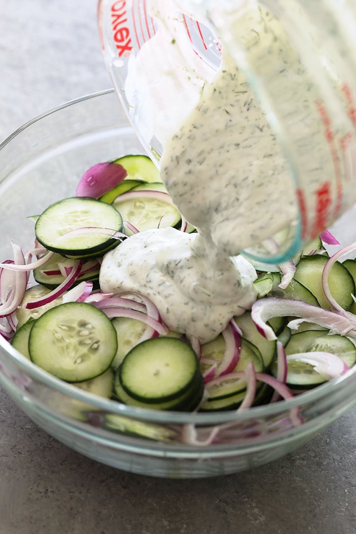 If you are looking for a healthy side salad for your next BBQ or a healthy salad for meal prep, look no further! This creamy cucumber salad is simple, flavorful, and delicious. Pouring dressing on cucumber salad.