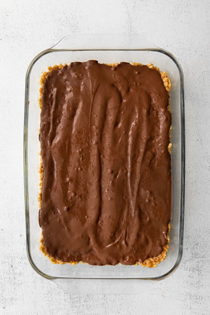 These healthy scotcheroos are made with all natural sugar, unsalted peanut butter, and coconut oil (no white sugar or corn syrup)! They are ready in no time and a great healthy spin on this classic dessert bar. melted chocolate layer of healthy scotcheroos in a baking dish